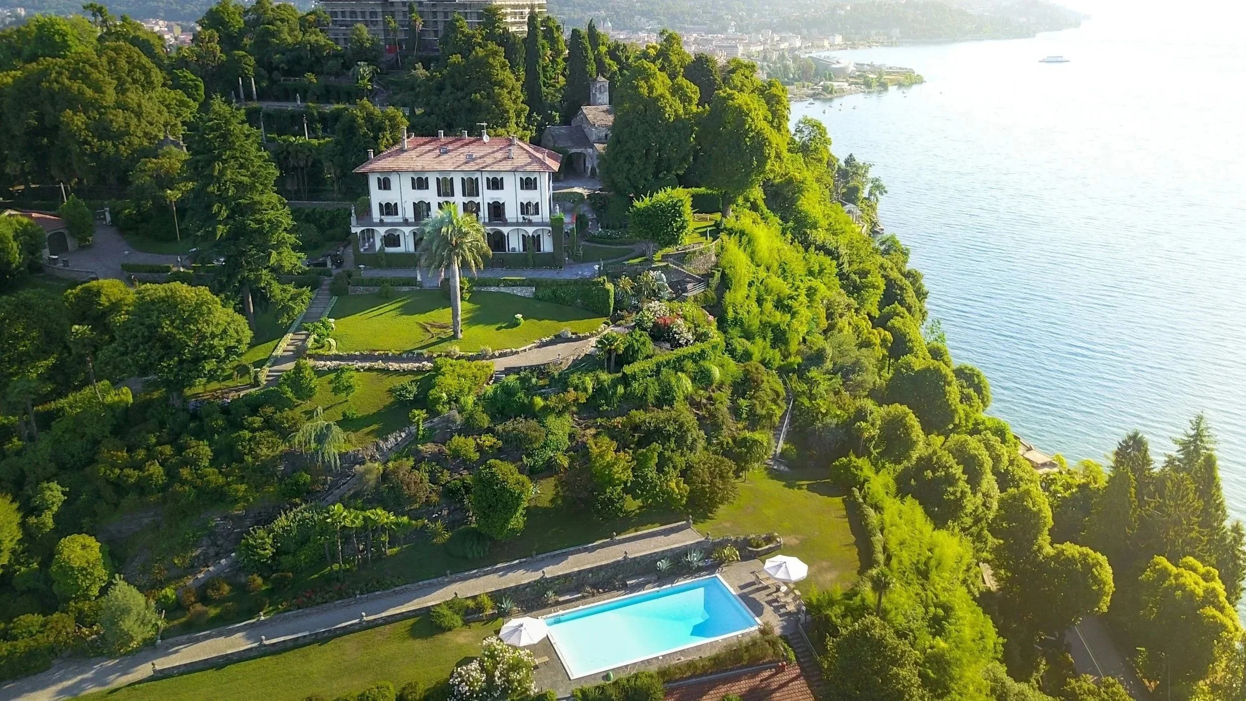 Aerial view of a luxury house on a lush, green hillside beside a body of water, with a swimming pool, gardens, and dense trees surrounding the property.
