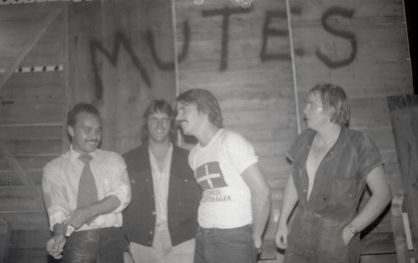 Four people in a wooden room, one man and three women, are engaged in conversation. The man has a mustache, is wearing a white shirt, and is holding a drink. The woman to his left has his arm around her waist and is smiling, the second woman from the