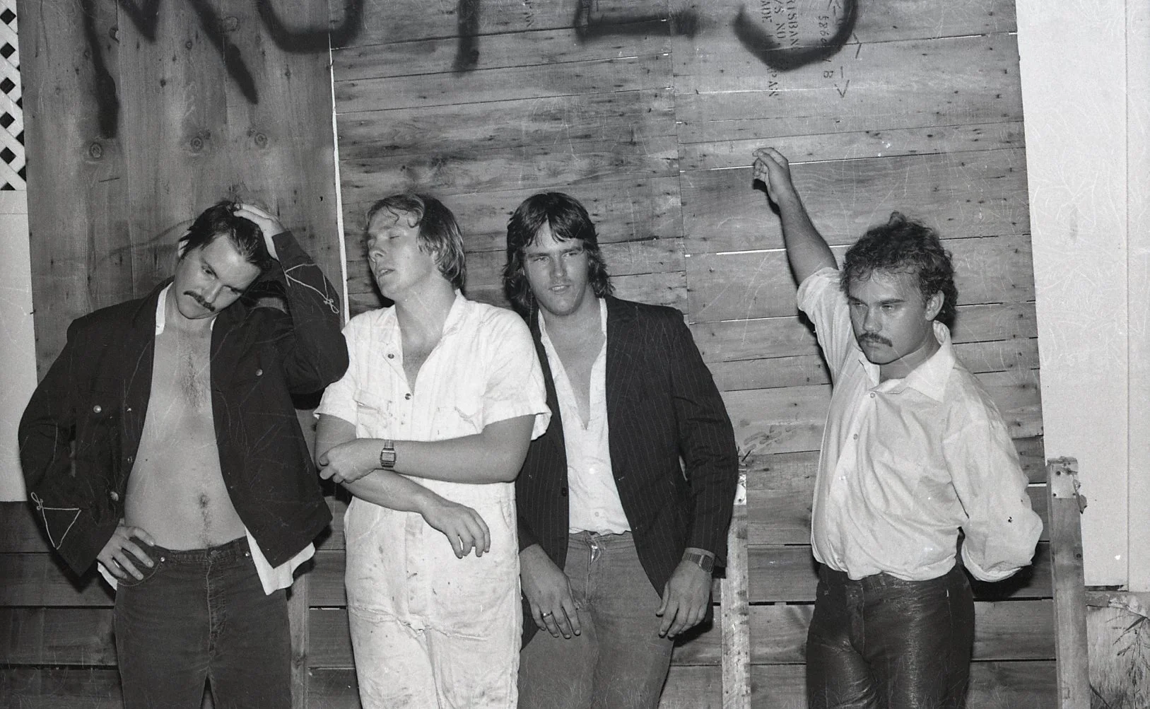 Black and white photo of four men standing in front of a wooden wall. The man on the far left has a mustache, is shirtless with an open jacket, and is touching his head. The second man is wearing a white shirt, has his eyes closed, and arms crossed. 