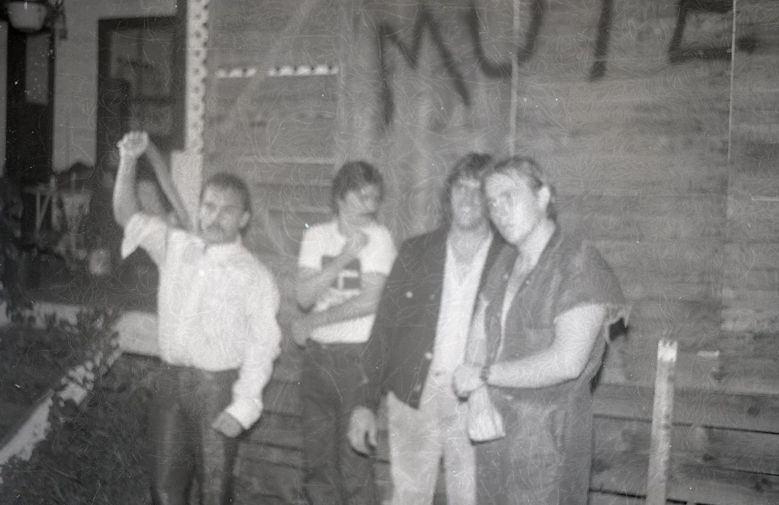 A black and white photo of four children standing outdoors, one girl on the far left raising her arms in the air, three boys in the middle and right, with one of them wearing glasses, in front of a wooden plywood wall.
