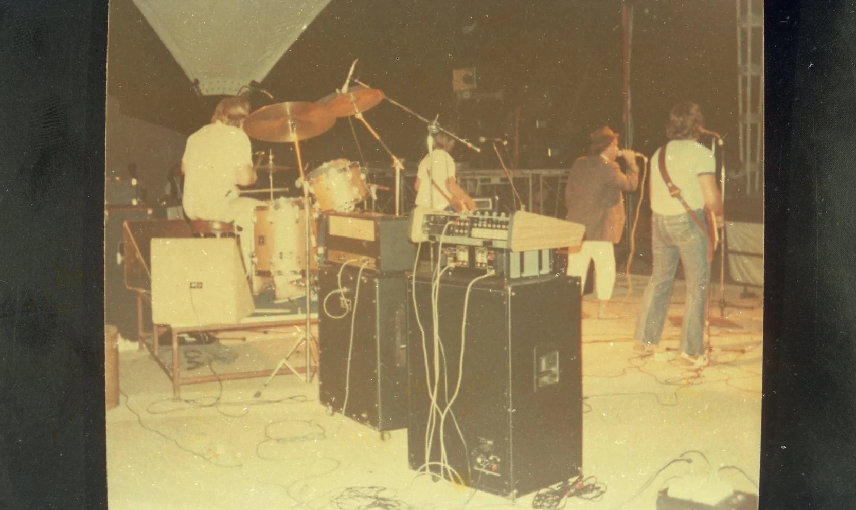 Four musicians performing on stage with various instruments, including drums, keyboard, and a guitar, during a live concert.