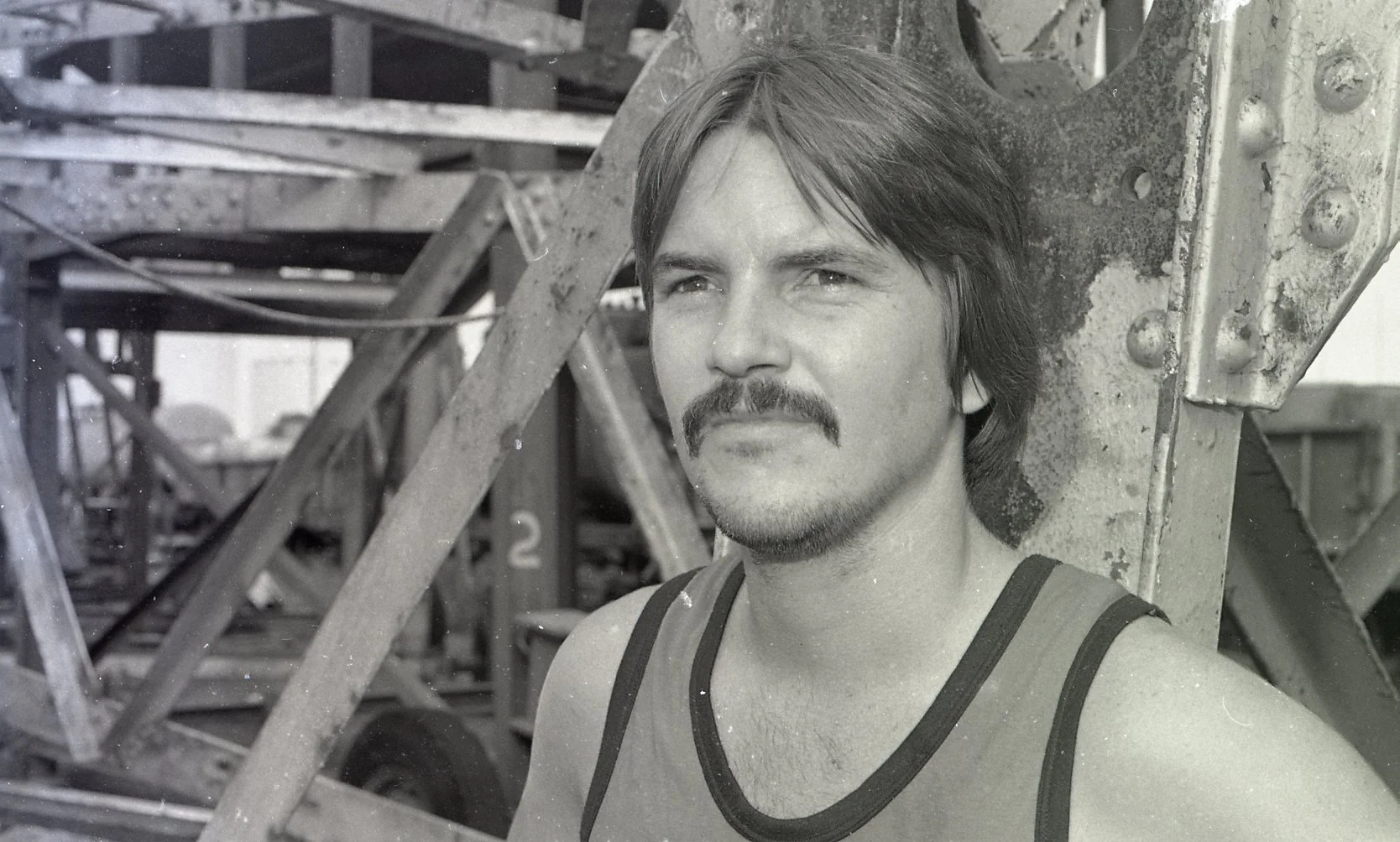 Black and white photo of a man with a mustache and short hair, wearing a tank top, leaning against a metal structure with an industrial background.