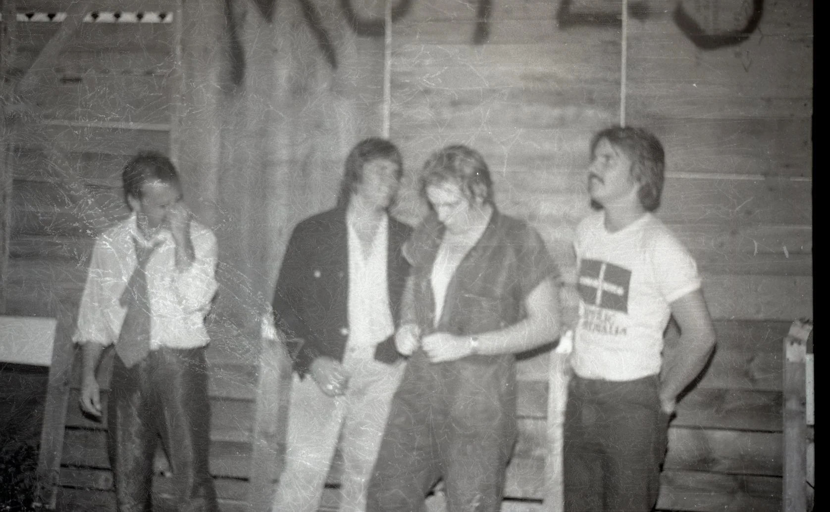 Black and white photo of four people standing indoors, two men are shaking hands, one person is wearing a shirt with a logo or emblem, and another is in a jacket. The background shows a wooden wall and a bicycle hangs on the wall.