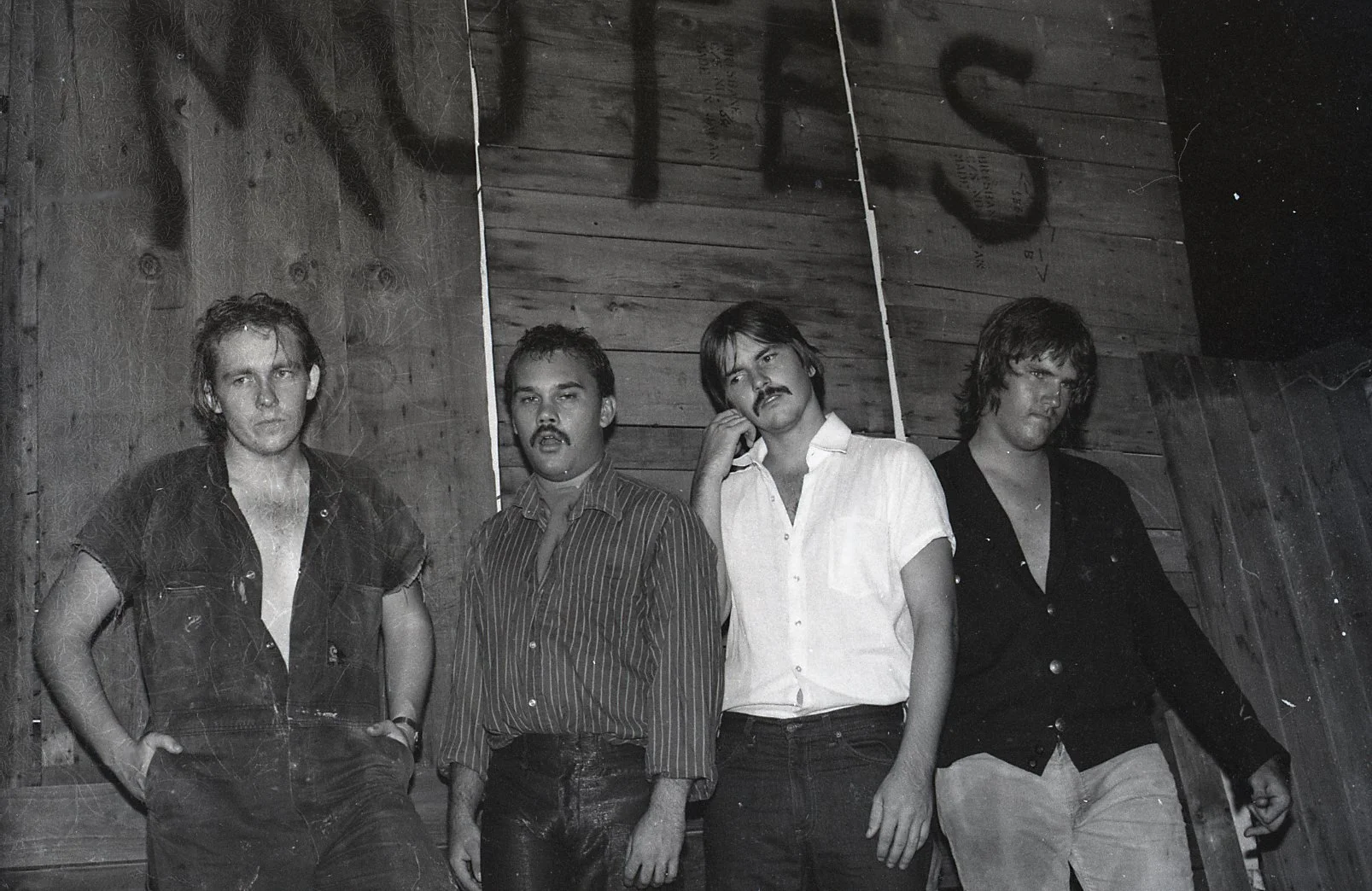 Black and white photo of four men standing in front of wooden walls, with some shadow messages written on the wall behind them.