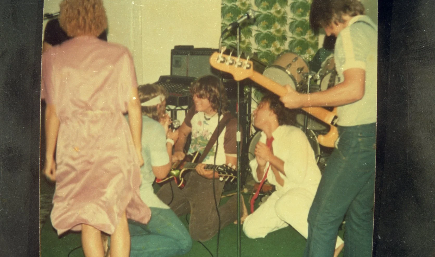 Group of children and teenagers engaging in a musical performance, with some playing guitars and drums, in a room with vintage-style wallpaper.