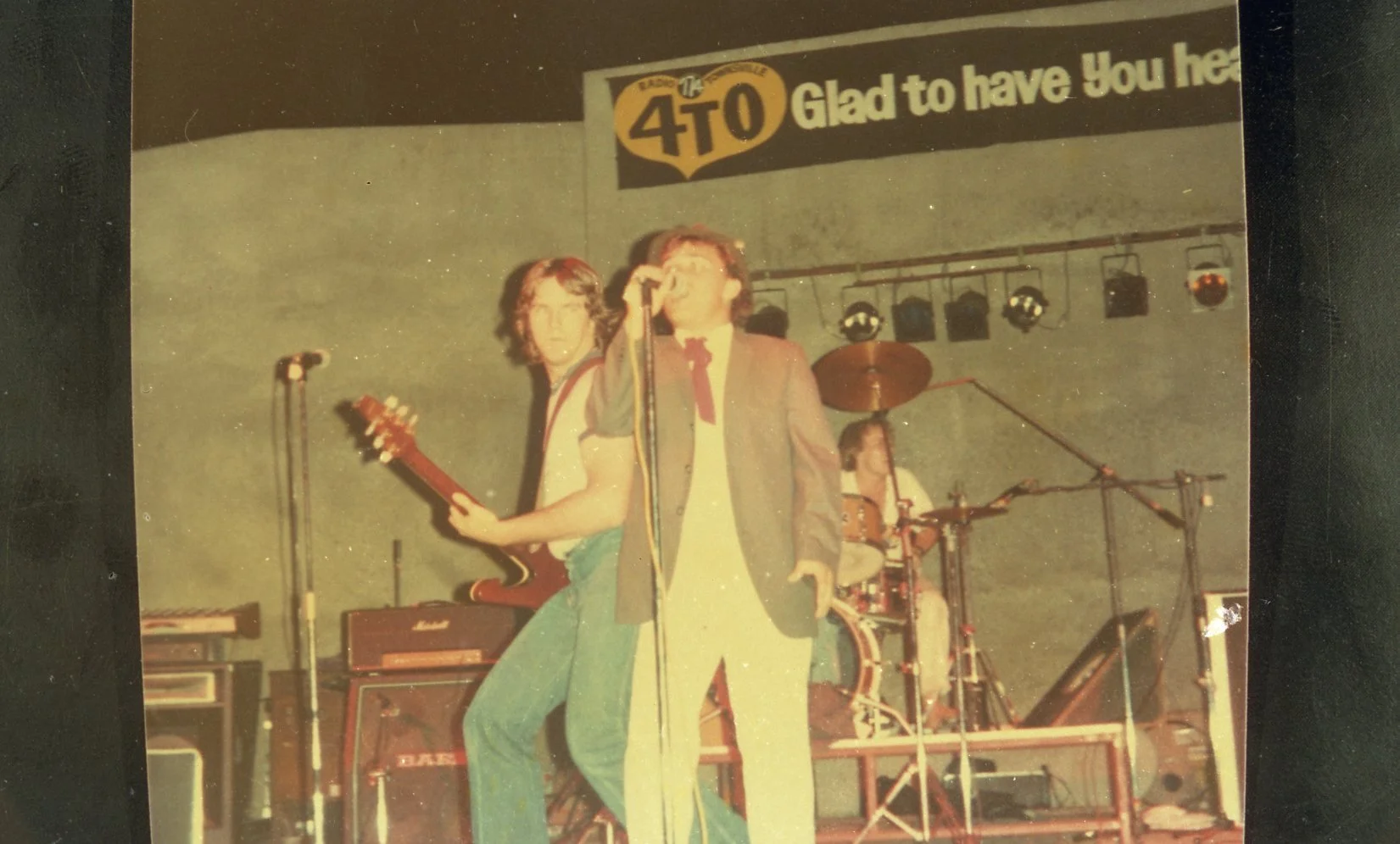 A band performing on stage with a guitarist, a singer, and a drummer in the background. The stage has a banner reading 'Glad to have You here' and the initials '4TO'.