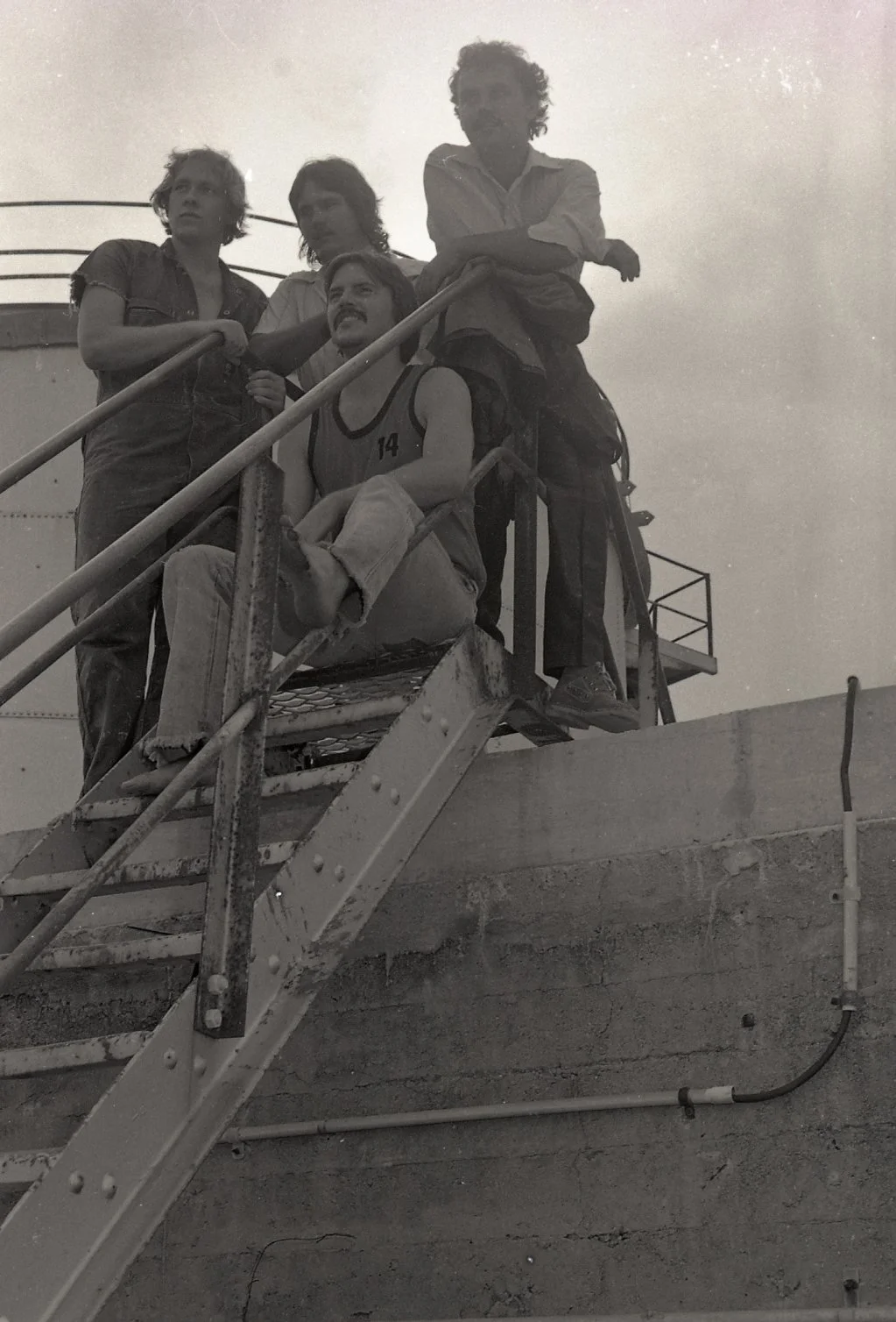 Four people, including one sitting, are on metal stairs outside a building, with two standing and leaning on the railing. The photo is black and white.