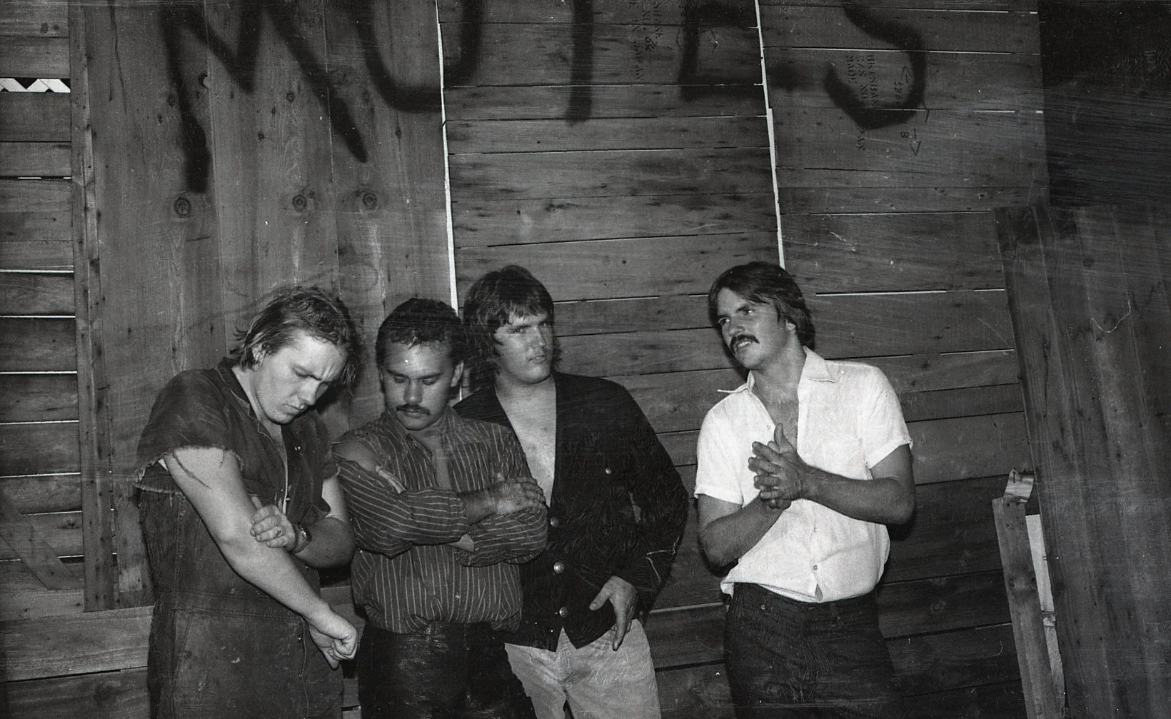 Black and white photo of four men standing against a wooden wall, with one man speaking and gesturing with his hands while the others listen.