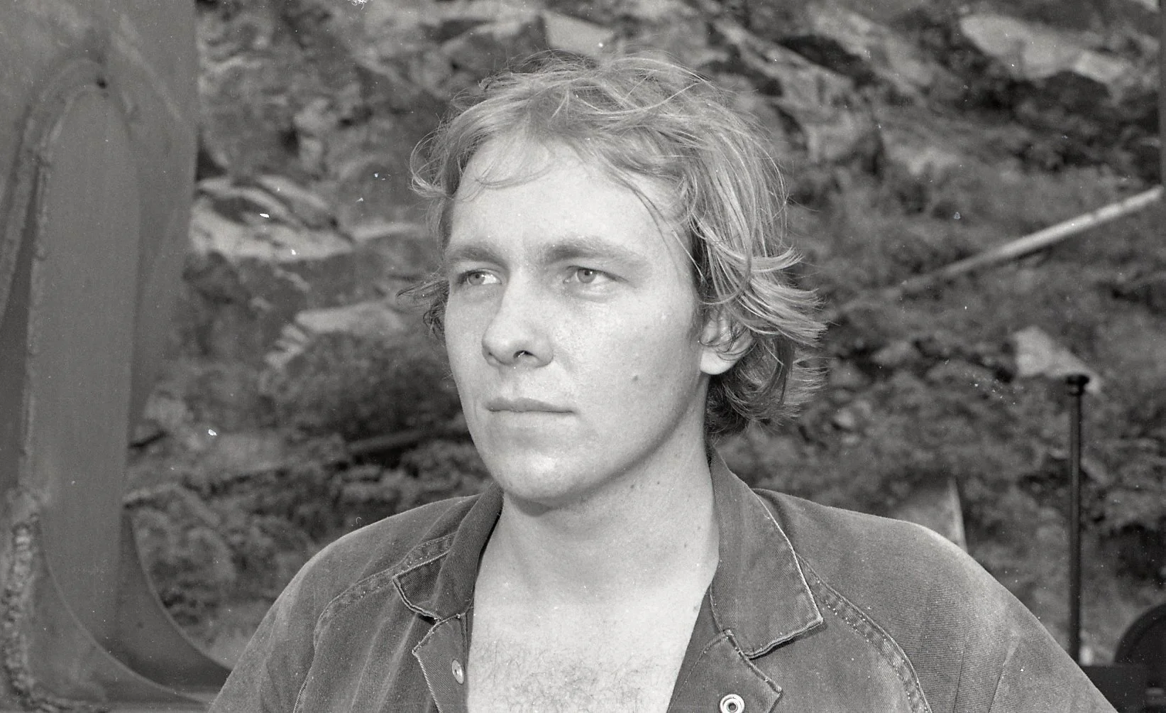 Black and white photo of a young man with wavy, medium-length hair, wearing a collared shirt, standing outdoors with a rocky background.