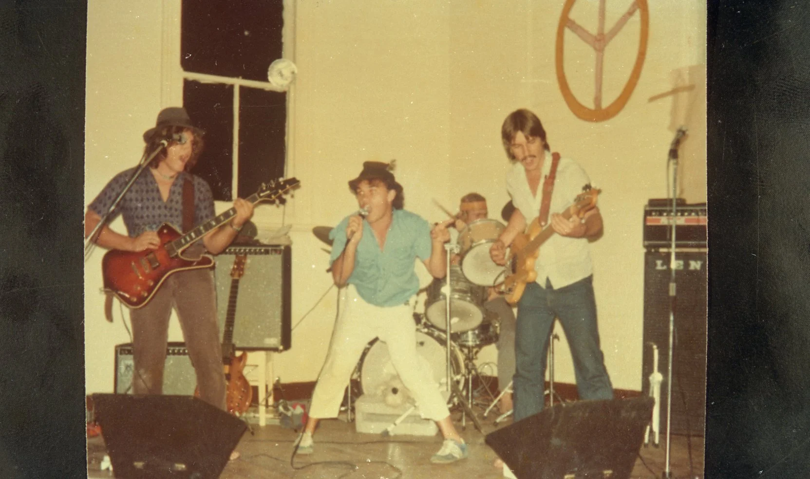 A band performing live on stage, with four members: a guitarist on the left, a singer in the center holding a microphone, a drummer behind the singer, and another guitarist on the right. The background has a peace symbol on the wall.