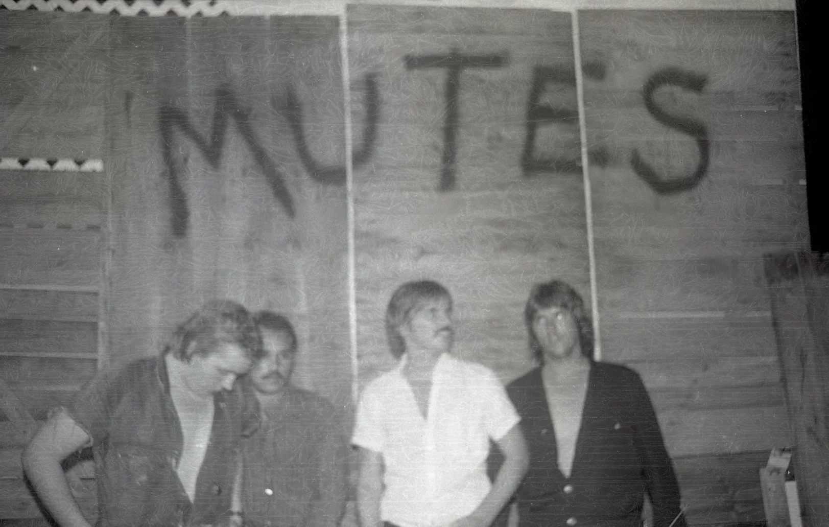 Four women standing indoors in front of a wooden wall. The woman on the left is looking down, wearing a vest. The second woman from the left has glasses and curly hair, smiling. The third woman from the left has short hair and is wearing a white shir