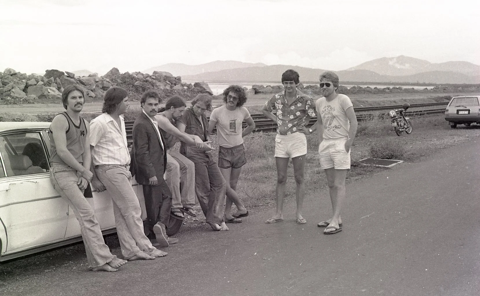 Black and white photo of eight men standing and leaning against a car by a railway track. They are outdoors in a rural area with mountains in the background. Some men are barefoot, wearing casual clothing, and one has sunglasses. A motorcycle and ano