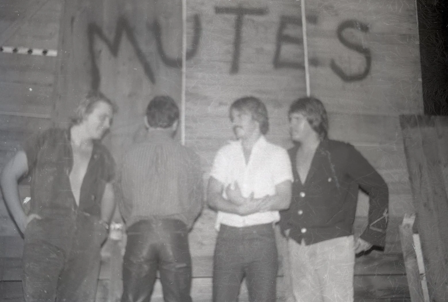 A black and white photo of four people standing indoors, with the word 'WETES' displayed on a banner behind them.