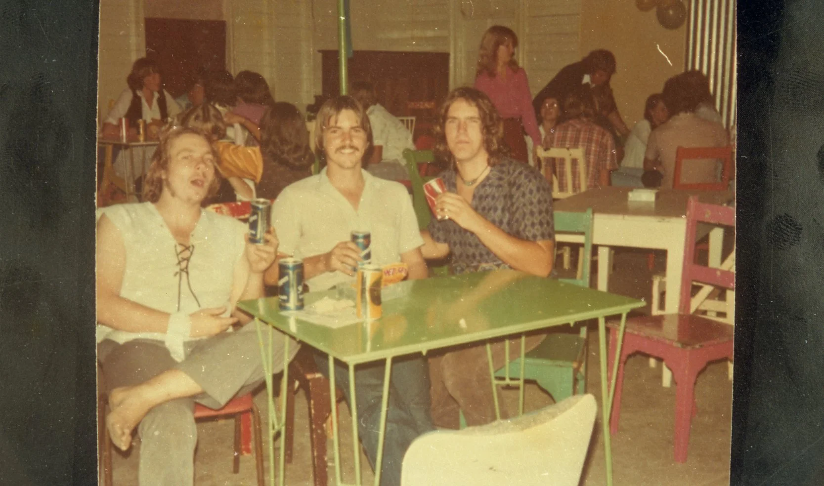 Three people sitting at a green table drinking soda at a social gathering, with other people milling around in the background.