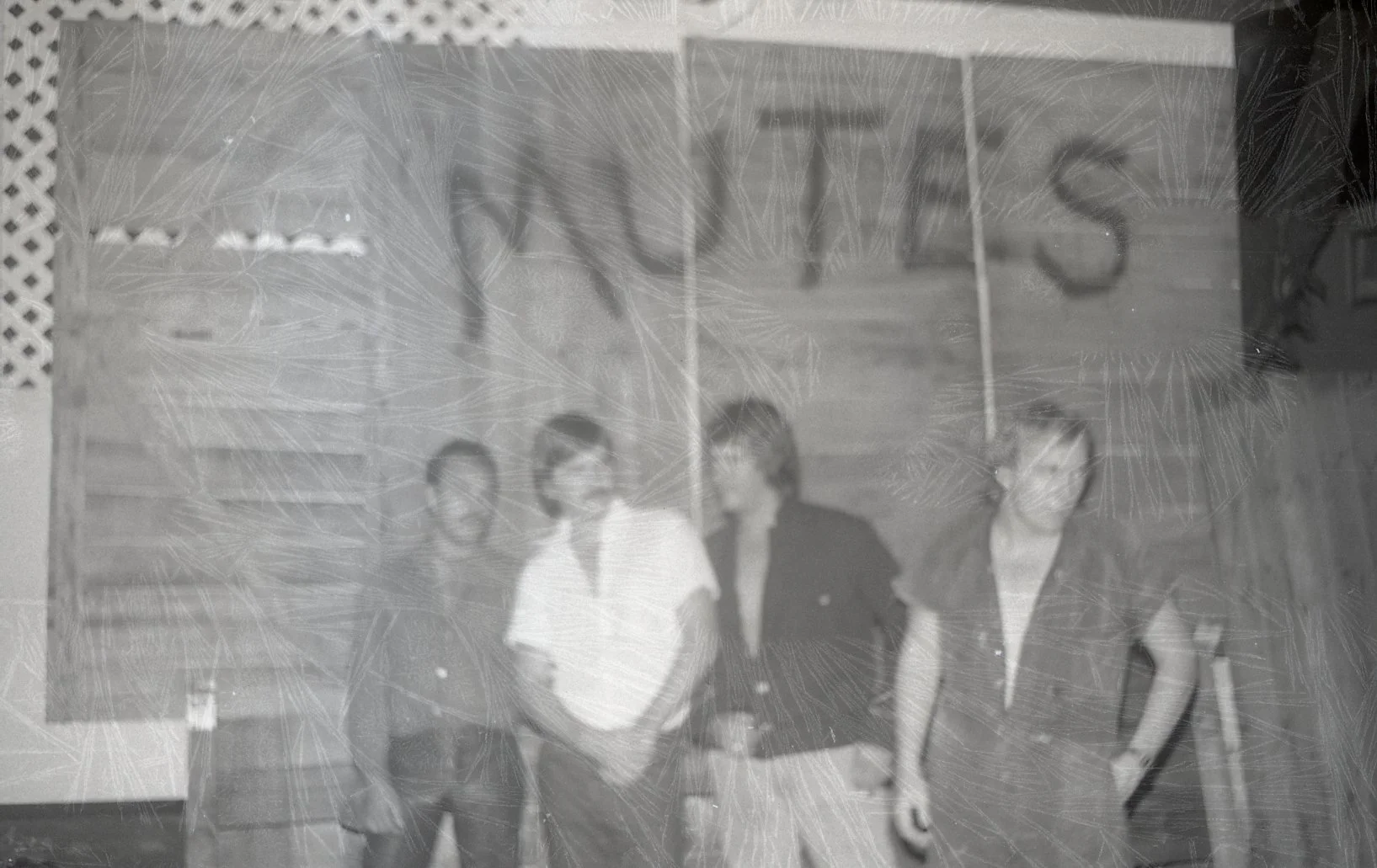 Black and white vintage photo of four people standing in front of a wooden building with stairs. The sign reading 'WHS' is visible above them.