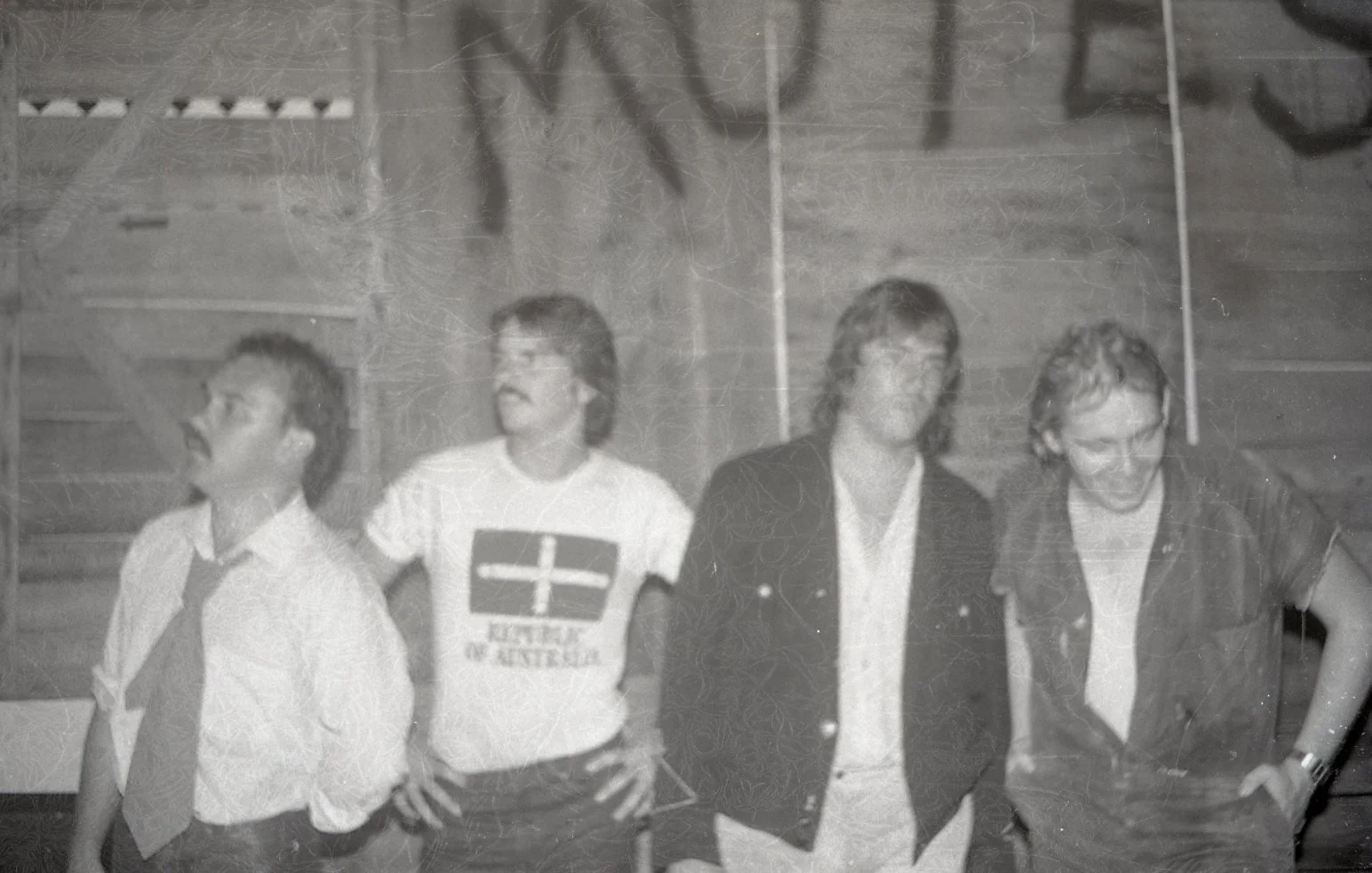 Four men standing side by side in a gymnasium with wooden bleachers in the background. The man on the far left is wearing a collared shirt and tie, the second man is wearing a T-shirt that reads 'Dutch in Australia', the third man is wearing a dark b
