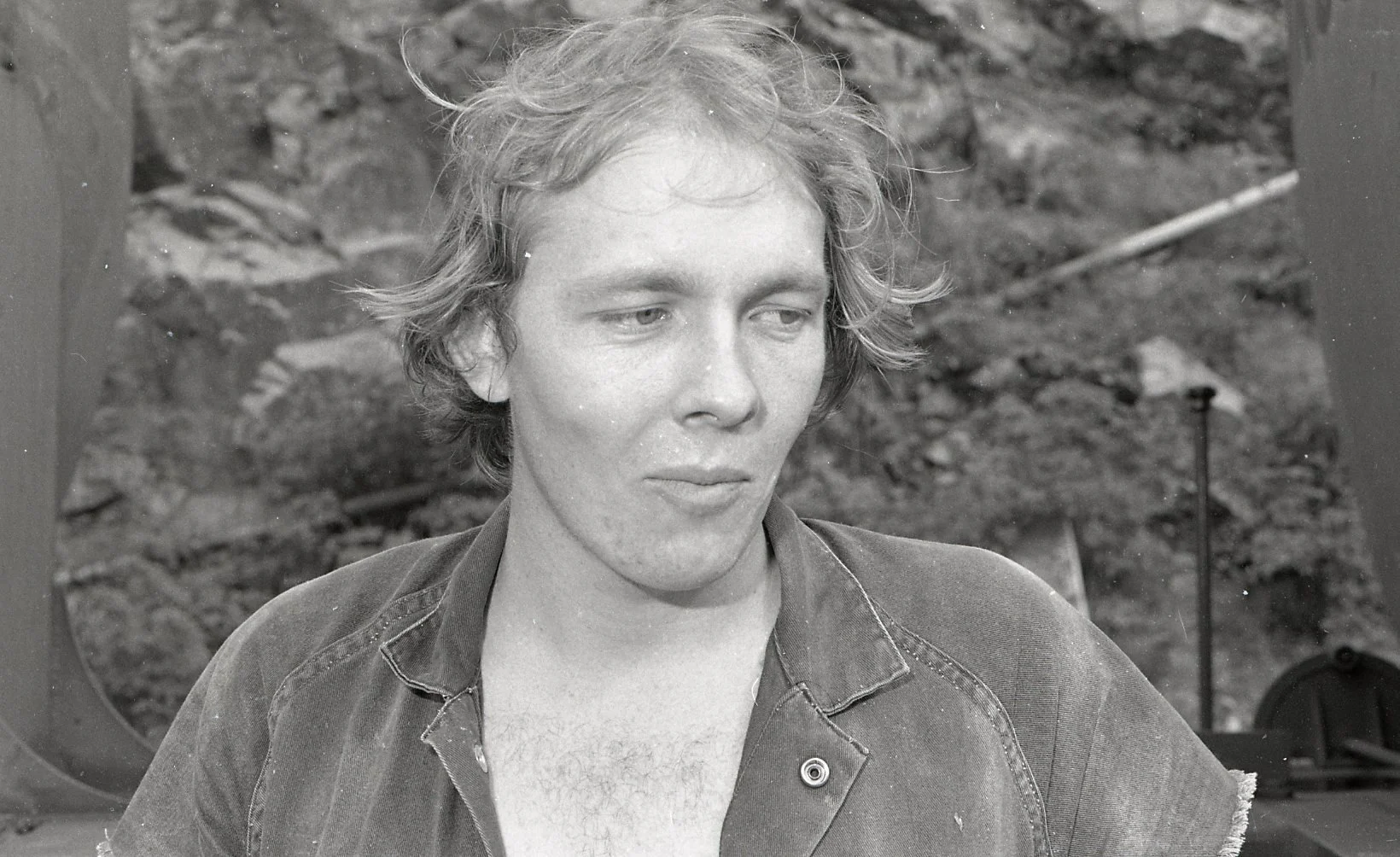 A young man with wavy hair and a denim jacket looking down, outdoors with rocky terrain in the background.