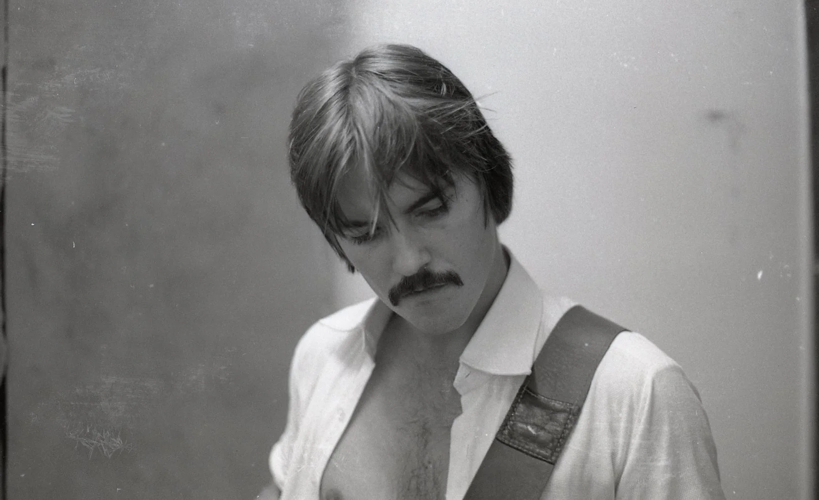 Black and white photo of a man with a mustache and disheveled hair, wearing an open shirt and carrying a guitar strap over his shoulder, looking downward.