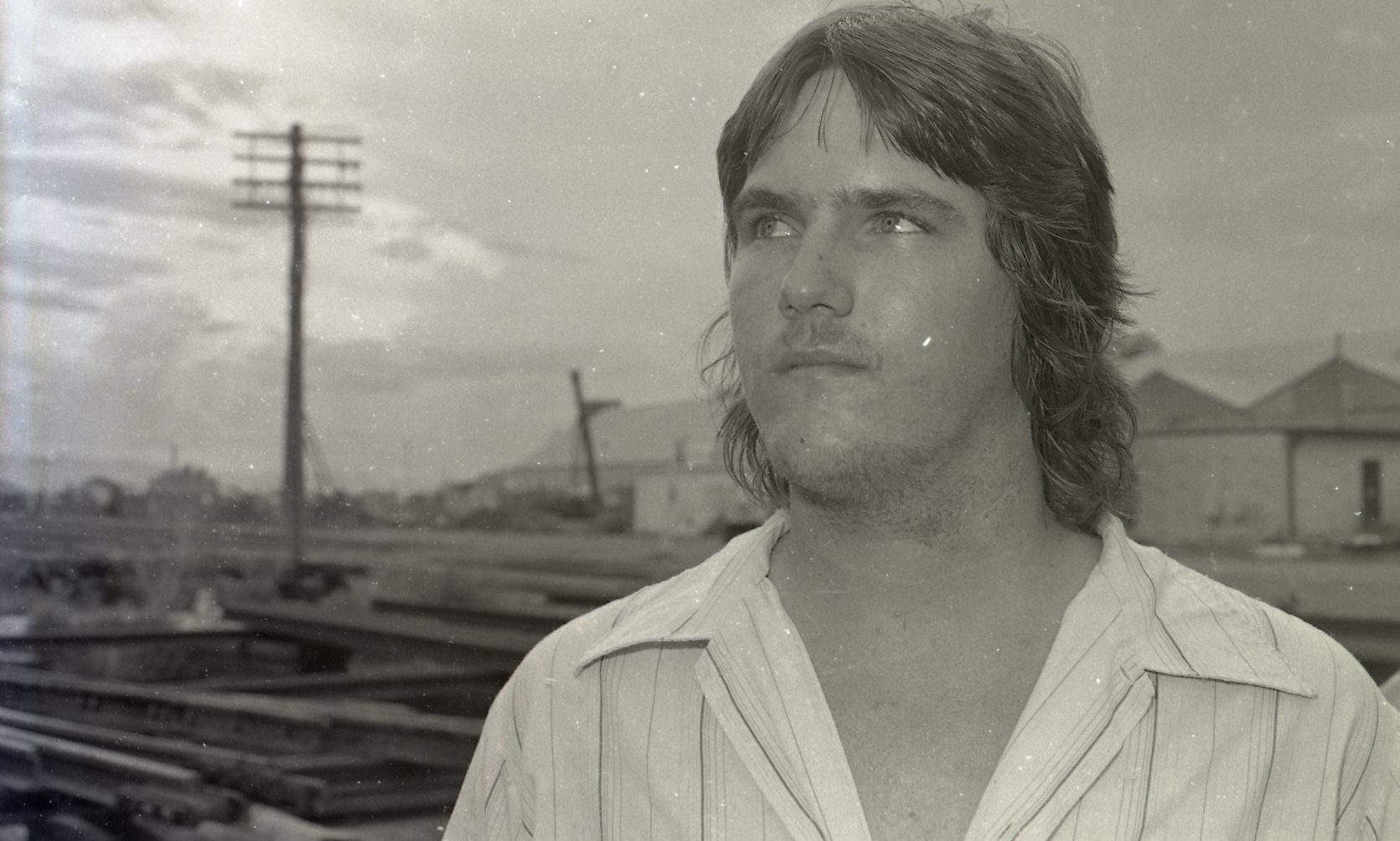 A black and white photo of a young man with medium-length hair, standing outdoors near train tracks. Wires and houses are visible in the background, and he is looking off to the side.