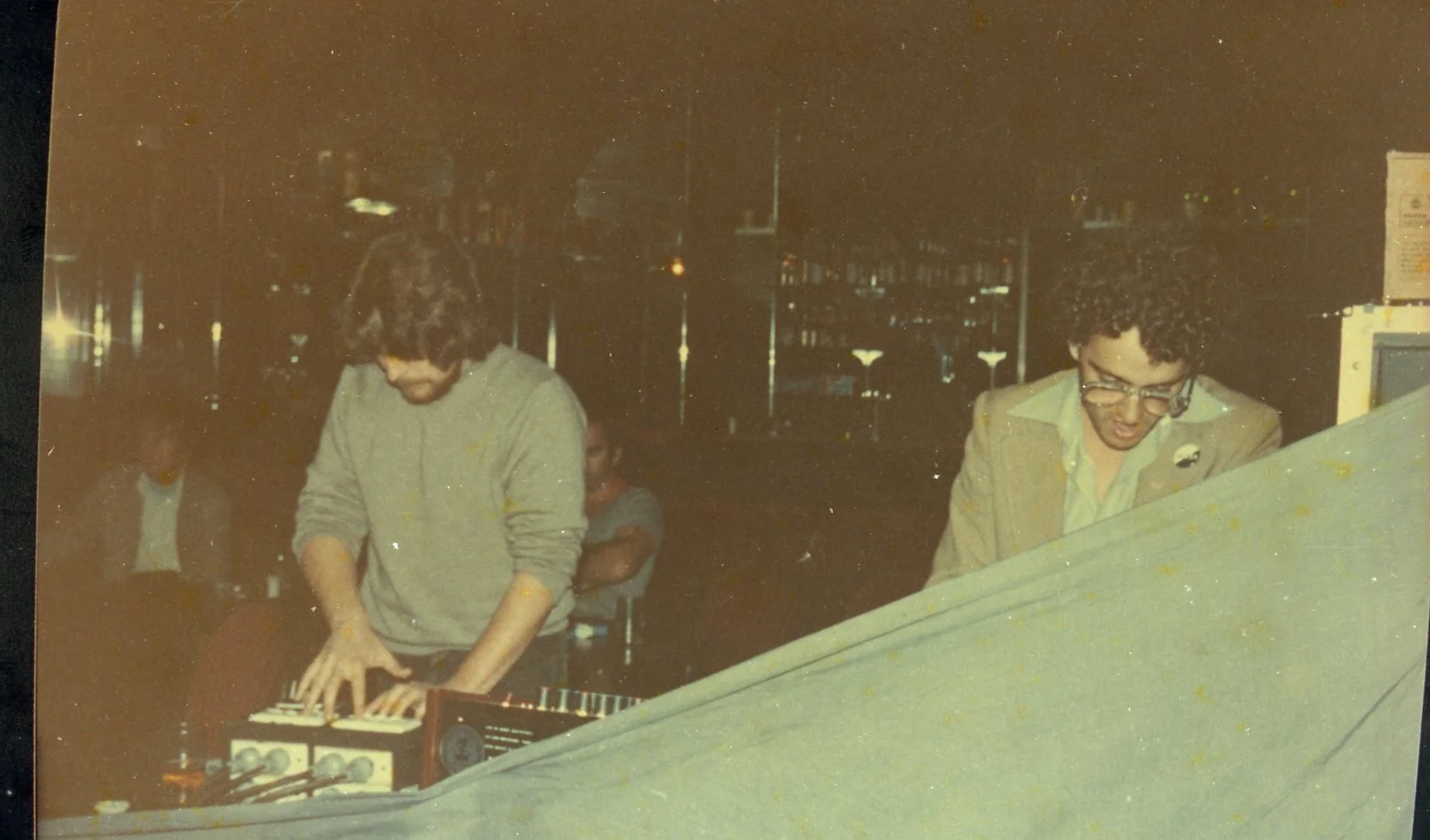 Two men with curly hair, wearing glasses and dressed in casual and business casual clothing, are playing electronic music instruments in a dimly lit indoor setting with a bar or cafe background. One is focused on a keyboard, the other on a mixing con