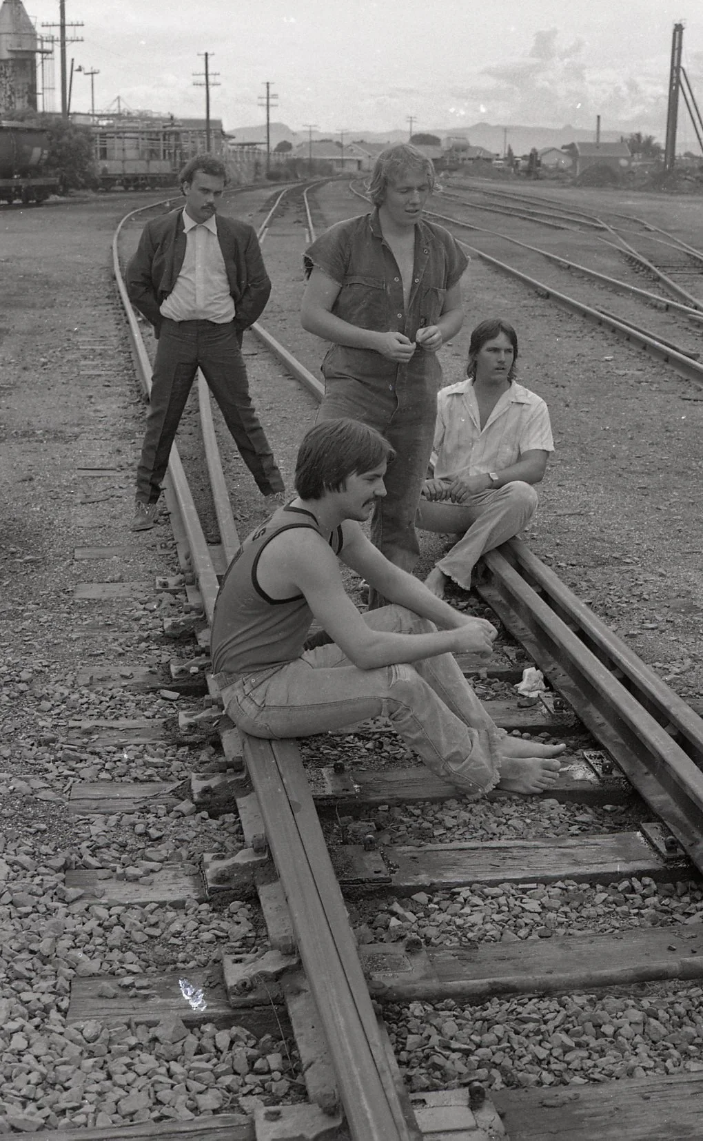 Four young adults sitting and standing on train tracks in an industrial area with train cars, power lines, and mountains in the background, during daytime.