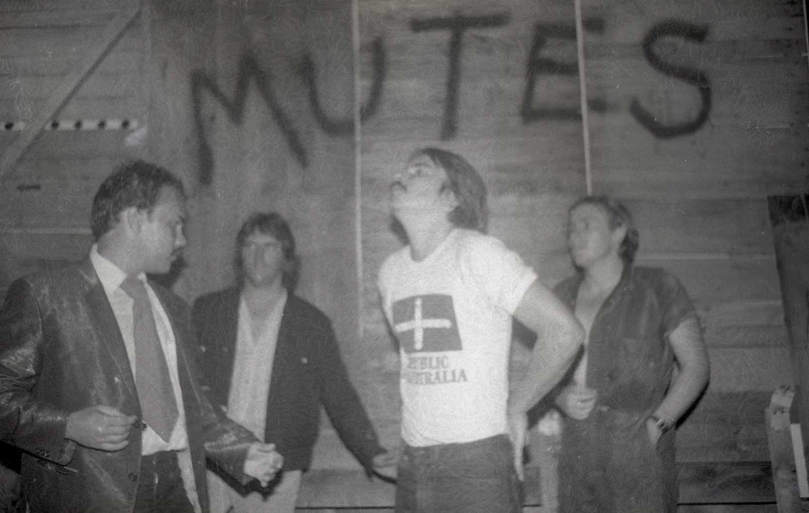 Black and white photo showing four people, three women and one man, standing inside a room with wooden panel walls. One woman is wearing a T-shirt with a cross and the words "EUC AUSTRALIA". The other woman is wearing a jacket. The man is wearing a s