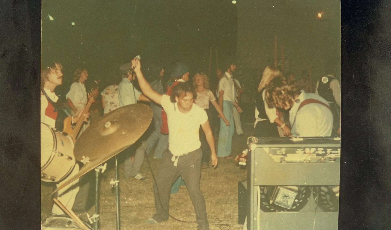 A vintage photo of a group of young people at a party or gathering, with some playing musical instruments like guitar and drums, and others dancing. The scene is indoors or perhaps outside at night with warm lighting.