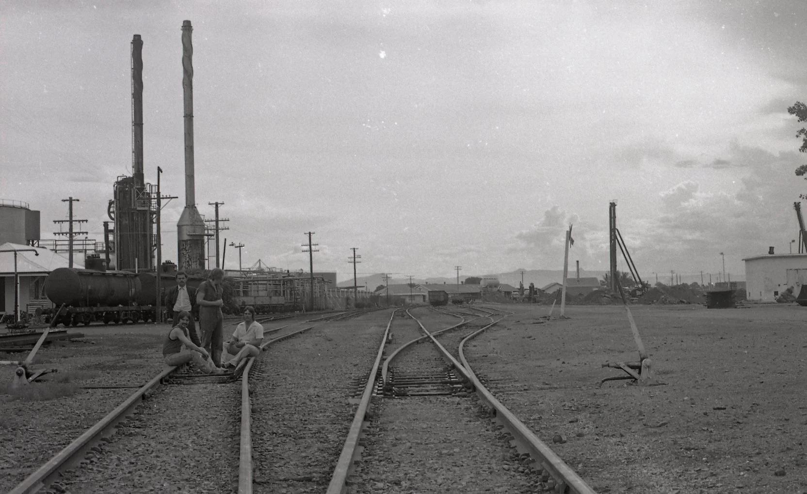 Black and white photo of a train yard with three people sitting and standing on the tracks, industrial buildings, utility poles, and distant mountains under a cloudy sky.