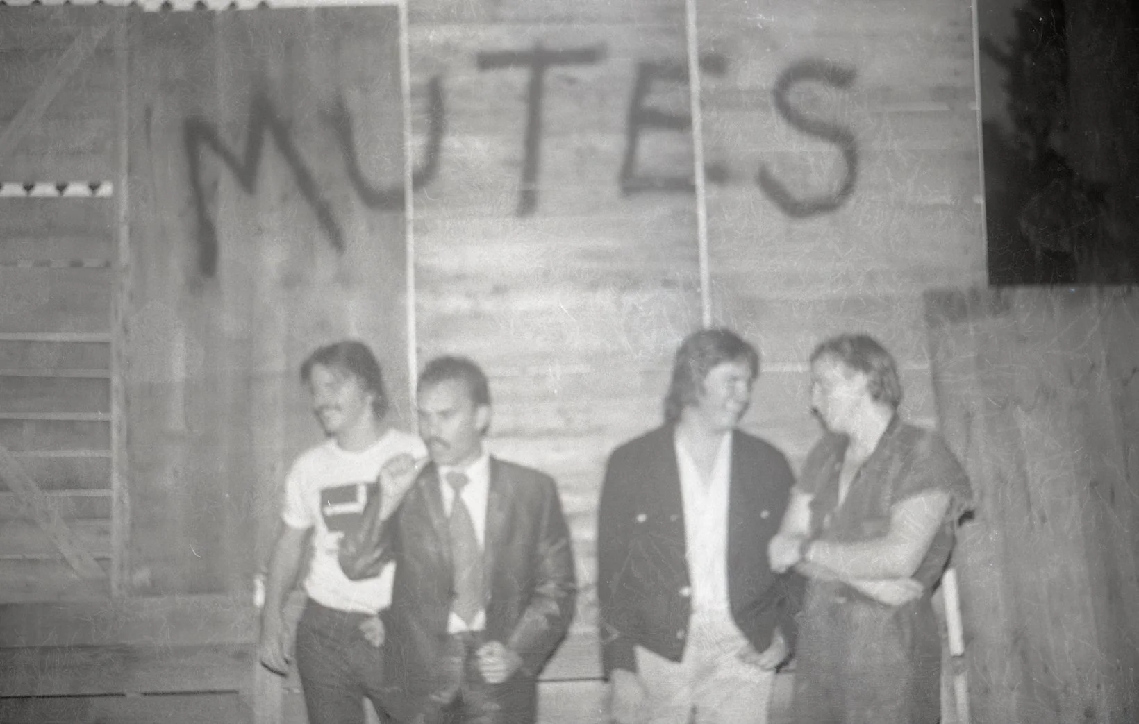 Four people standing against a wooden wall with the word 'WUTS' written above them. One person is holding a camera or phone, and the others are engaged in conversation.