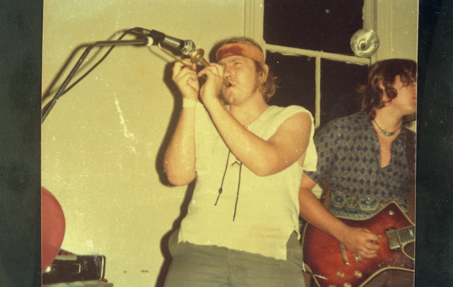 A young man with long hair, wearing a headband and a sleeveless shirt, is singing into a microphone during a performance. Another person with curly hair and a patterned shirt is playing an electric guitar in the background.