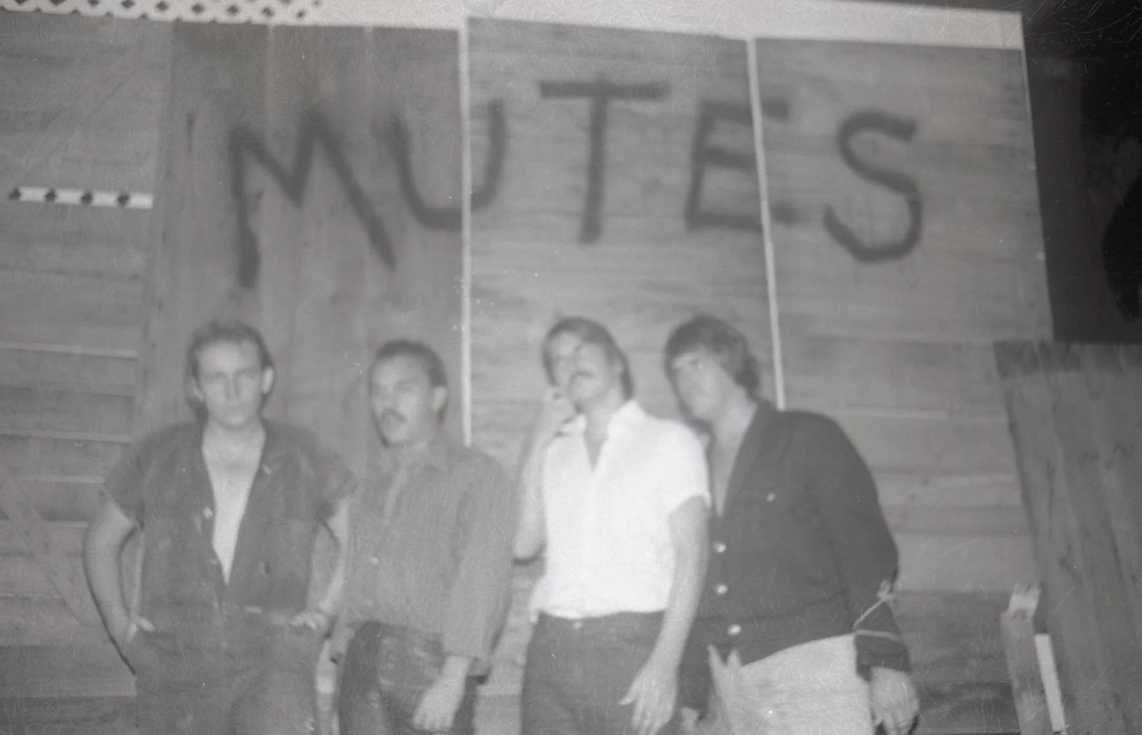Four young men standing in front of a wooden wall with a large sign that reads 'WUTES.'