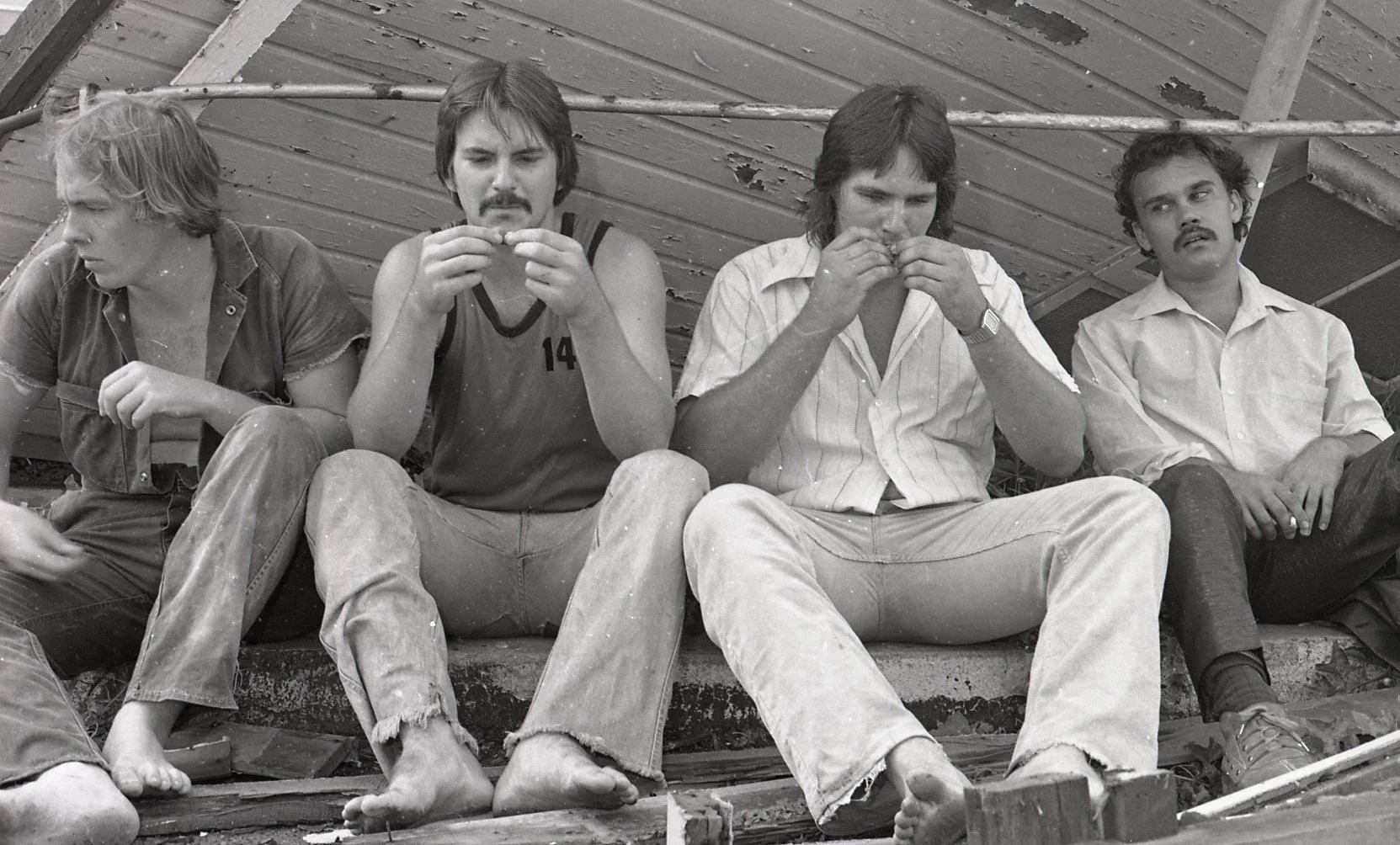 Four men sit on a concrete ledge in front of a weathered wood wall, with some construction materials and debris around.