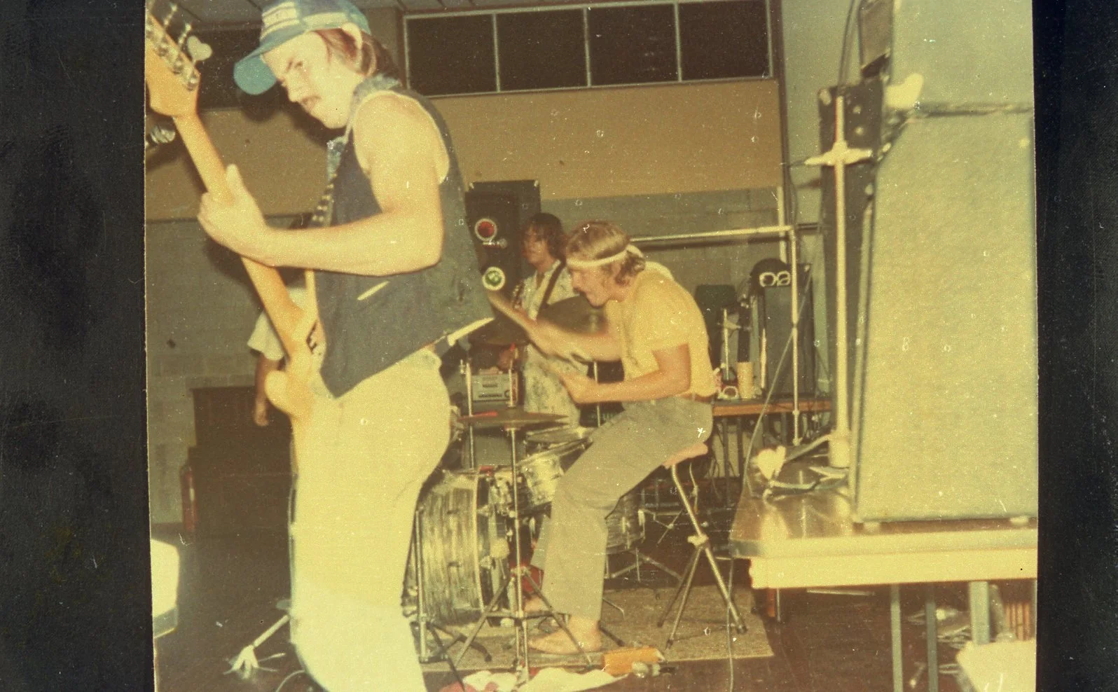 Three young men playing music on a stage, one with a bass guitar, another with a drum set, and a third with a microphone, in a room with equipment and shelves in the background, likely during a band rehearsal or performance.