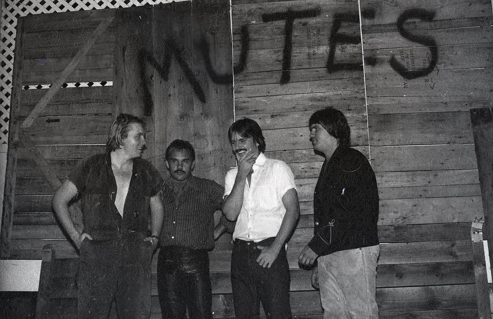 Four men are standing in front of a wooden wall with the word 'NUEVO' spray-painted on it. They are engaged in a conversation, with one man smiling and touching his chin, two men looking at each other, and one man looking at the camera. The setting a