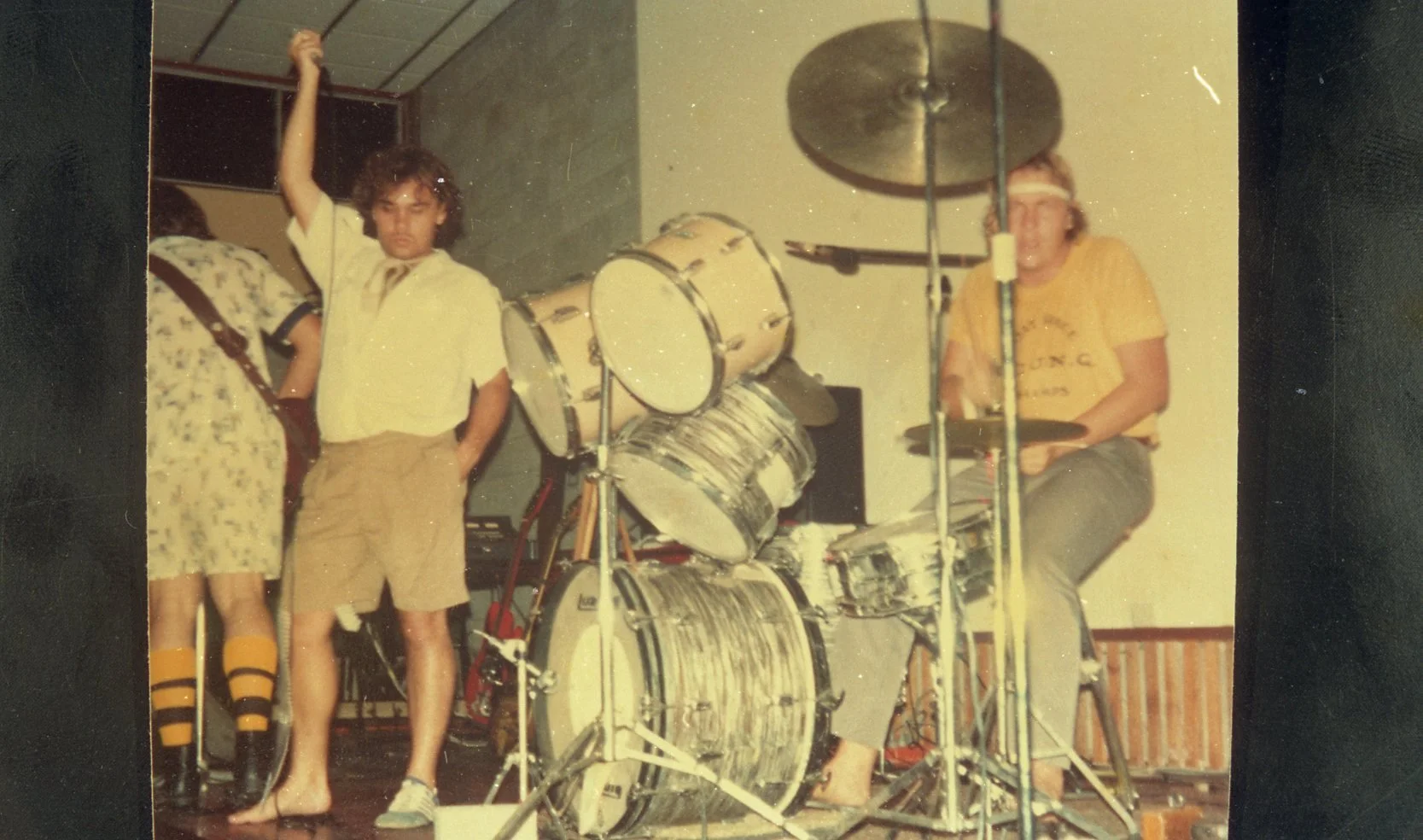 A vintage photo of a group of young people with a drum set and other musical equipment in a room, possibly for a band practice or performance.
