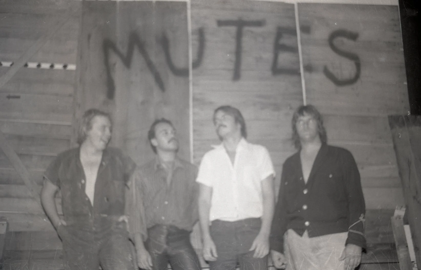 Black and white photo of four people standing in front of wooden panel walls. The letters mantra spelled out above their heads.