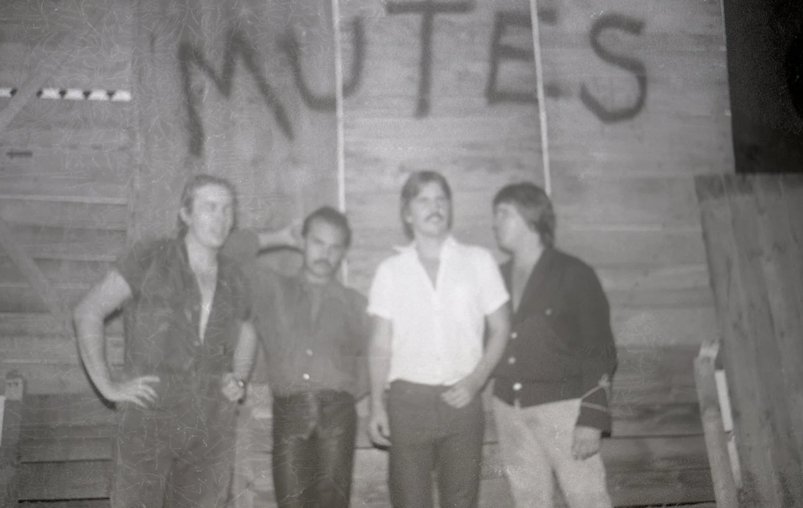 Black-and-white photo of four people standing together in front of a wooden wall with the letters "NPS" painted above them.