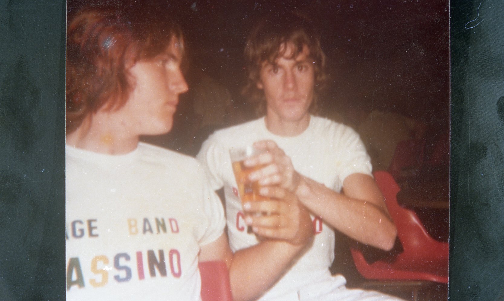 Two young men with shaggy hair in white t-shirts sitting in theater seats, one holding a glass of beer, with dark background.