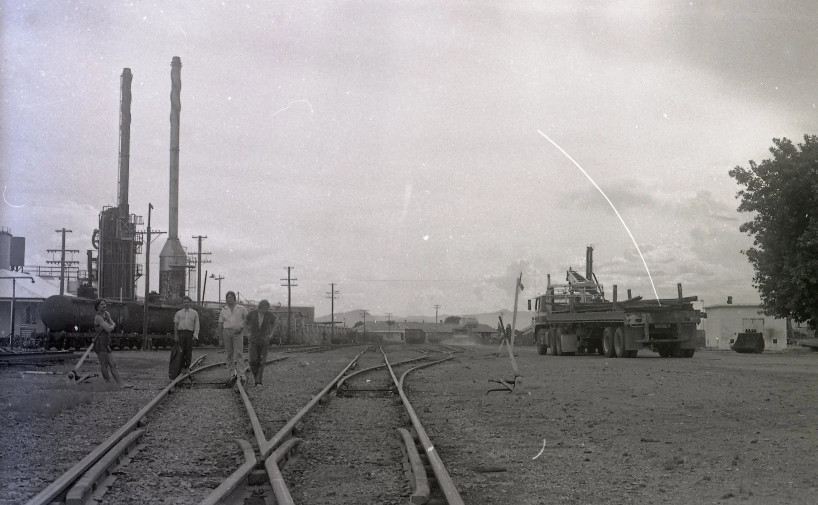 Black and white photograph of three people walking on railroad tracks near industrial buildings, large tanks, and utility poles, with a truck parked to the right and a tree on the far right.
