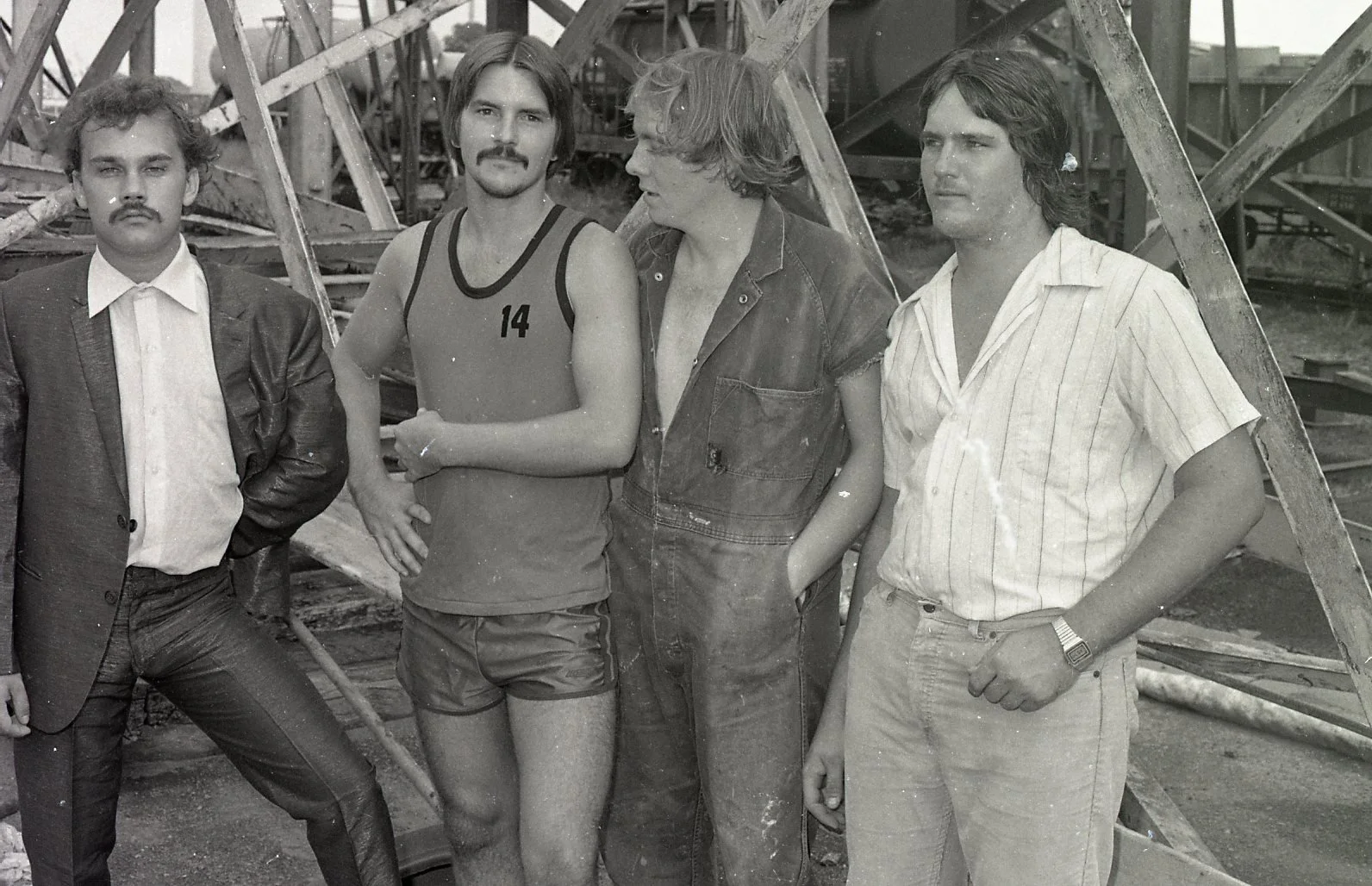 Four men standing outdoors in front of a roller coaster, dressed in various casual and formal clothing, with one man in a tennis uniform, in black and white.