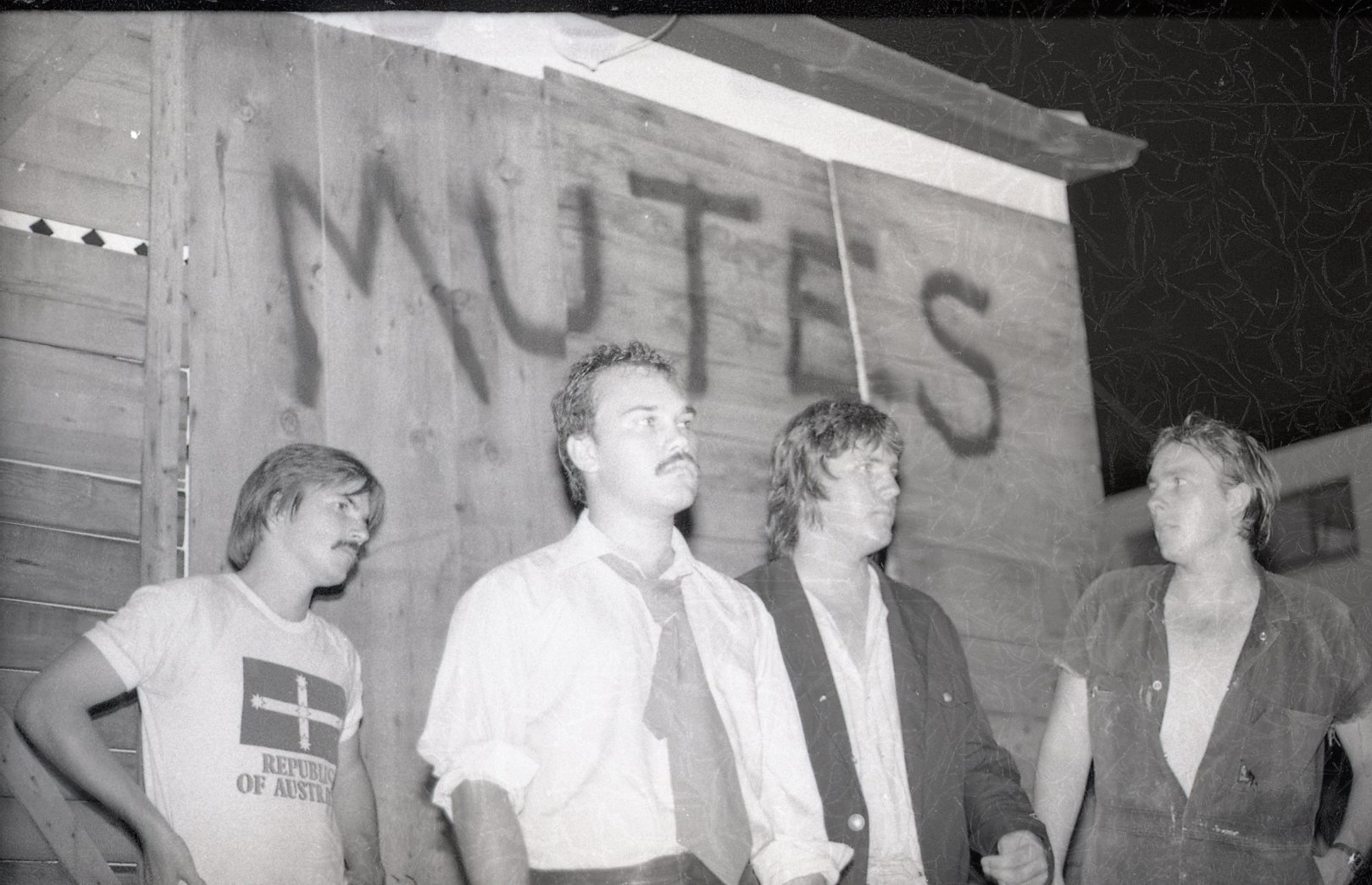 Four men standing in front of a wooden wall with the word "MYTTE" projected onto it. The man on the far left wears a t-shirt with a symbol and text, the second man from the left wears a shirt and tie, the third man wears a jacket over a shirt, and th