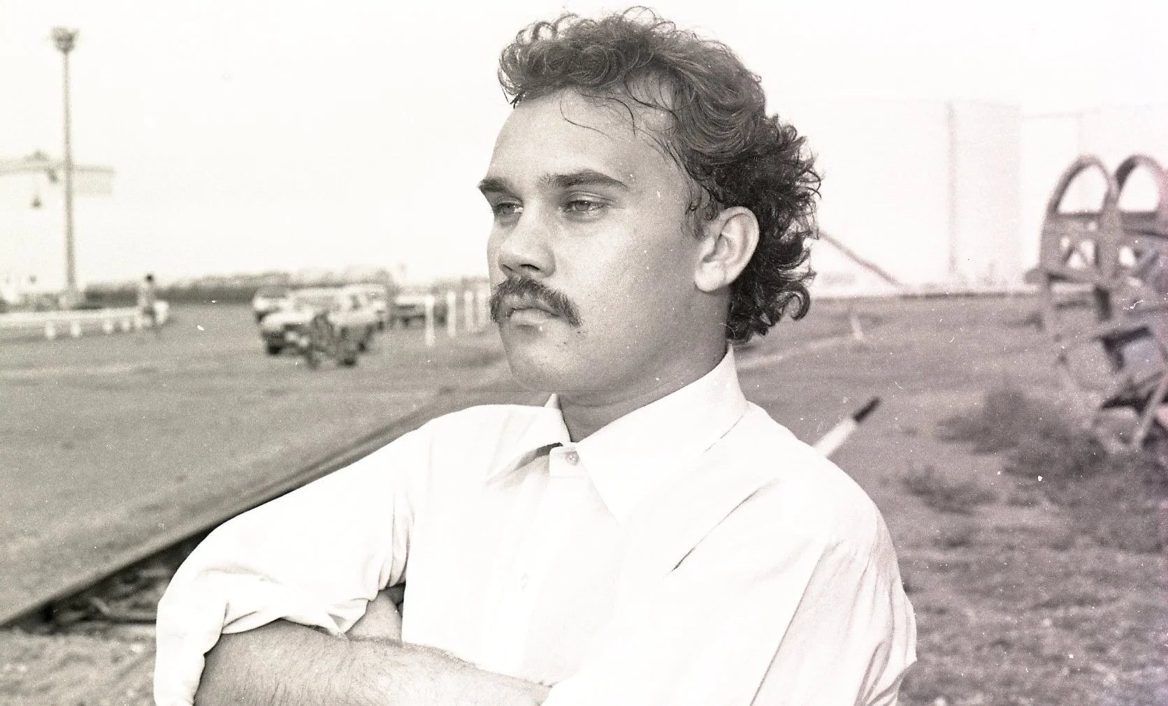 Black and white photo of a man with curly hair, mustache, and a serious expression, standing outdoors with arms crossed, in front of a blurry background of cars and structures.