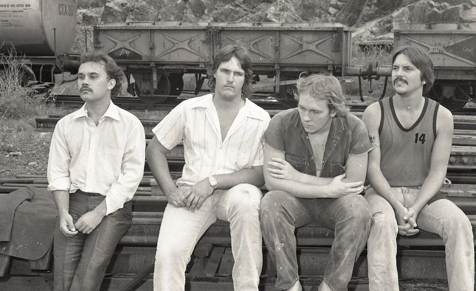 Black and white photo of four men sitting on train tracks with train cars in the background. All are casually dressed, with the man on the far right wearing a sports jersey numbered 14. They appear to be in a relaxed, contemplative mood.