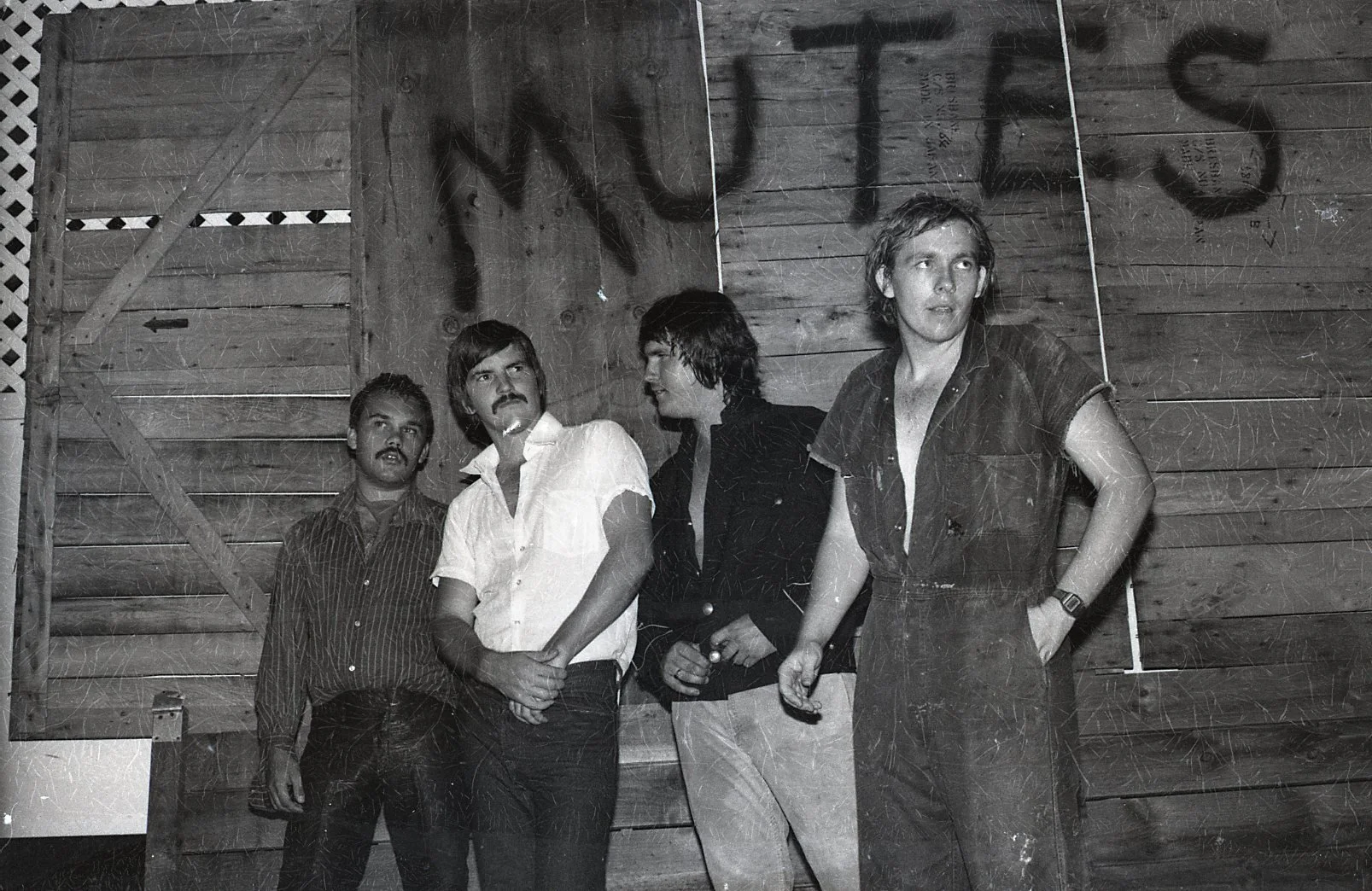 Black and white photo of four men standing against a wooden wall with the word "JUST" painted above their heads.