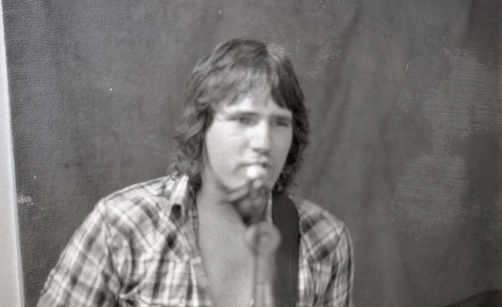 Black and white photo of a young man with shoulder-length wavy hair, wearing a plaid shirt, sitting against a textured wall. He appears to be in thought with a serious expression.