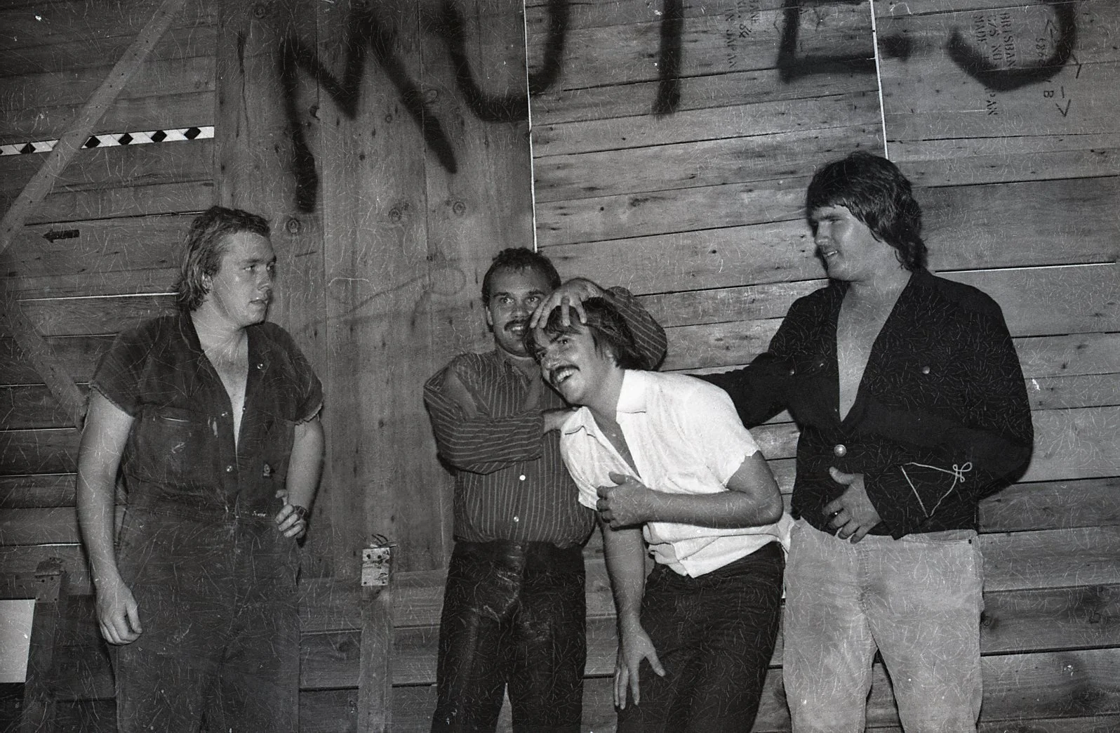 Four men, two with long hair, are in a black and white photo with a wooden background. One man is pinching the other man's nose, and the others are looking on. They appear to be having fun.