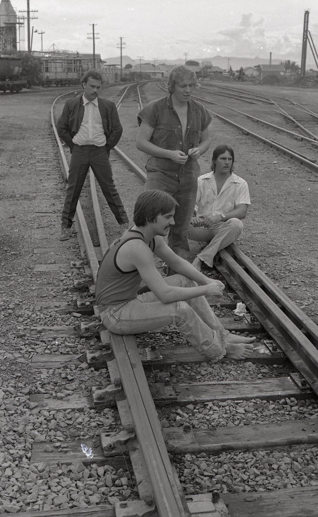 Four young people on a railroad track in an urban area, two sitting on the ground, one standing, and one walking, with shipping containers and utility poles in the background.