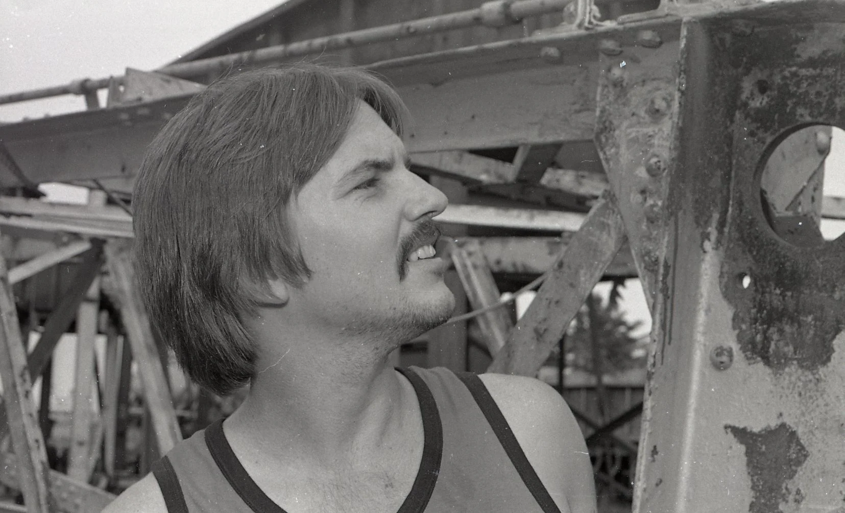 A young man with medium-length hair and a mustache, wearing a tank top, looking up at an industrial metal structure or machinery in an outdoor setting.