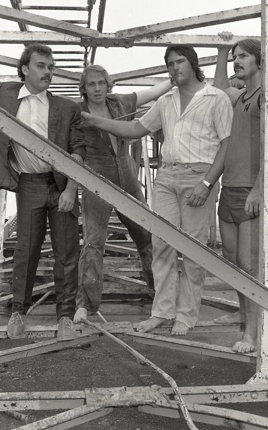 Black and white photo of four men standing on a construction site, surrounded by wooden scaffolding. They are dressed casually, with one in a suit, others in t-shirts and pants, and one barefoot. They are posing for the photo with serious expressions