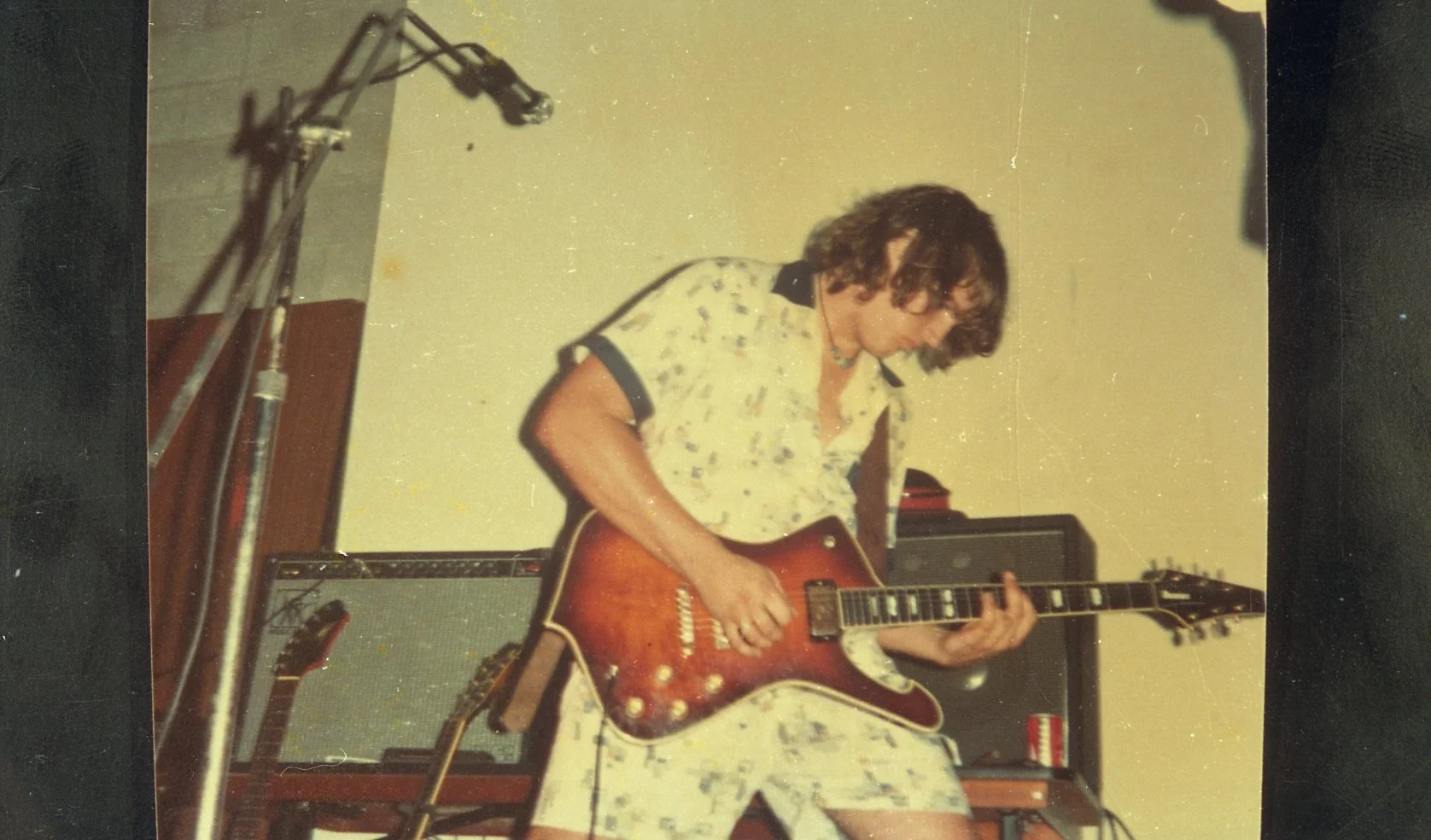 A person with curly hair playing an electric guitar in a room with musical equipment.